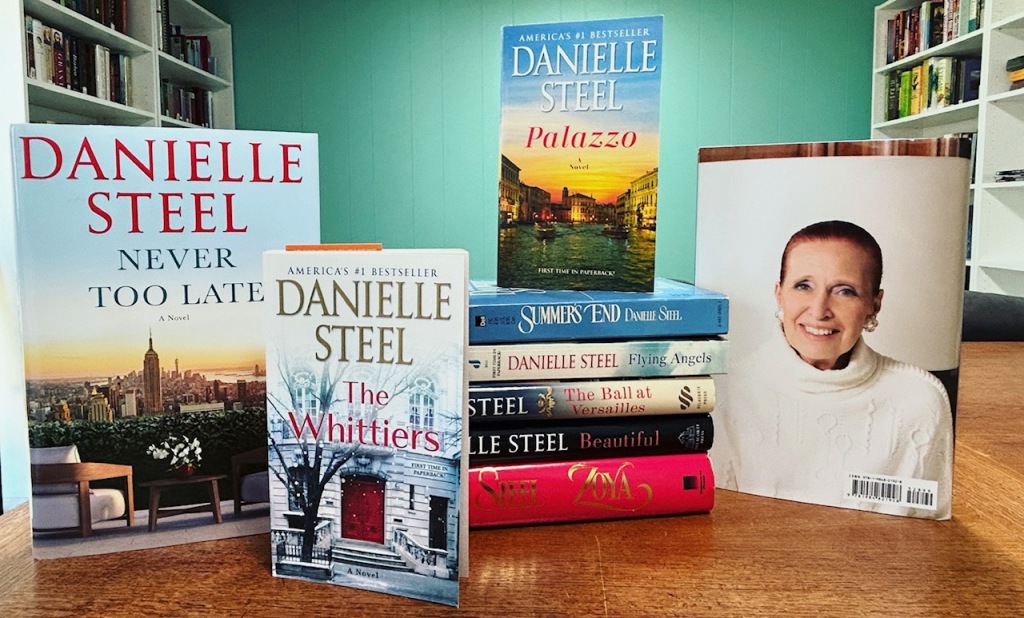 A group of book by author Danielle Steel sit artfully arranged on a wooden table. A photo of Danielle Steel, is also in the frame. There are white bookcases and a teal wall in the background of the photo.